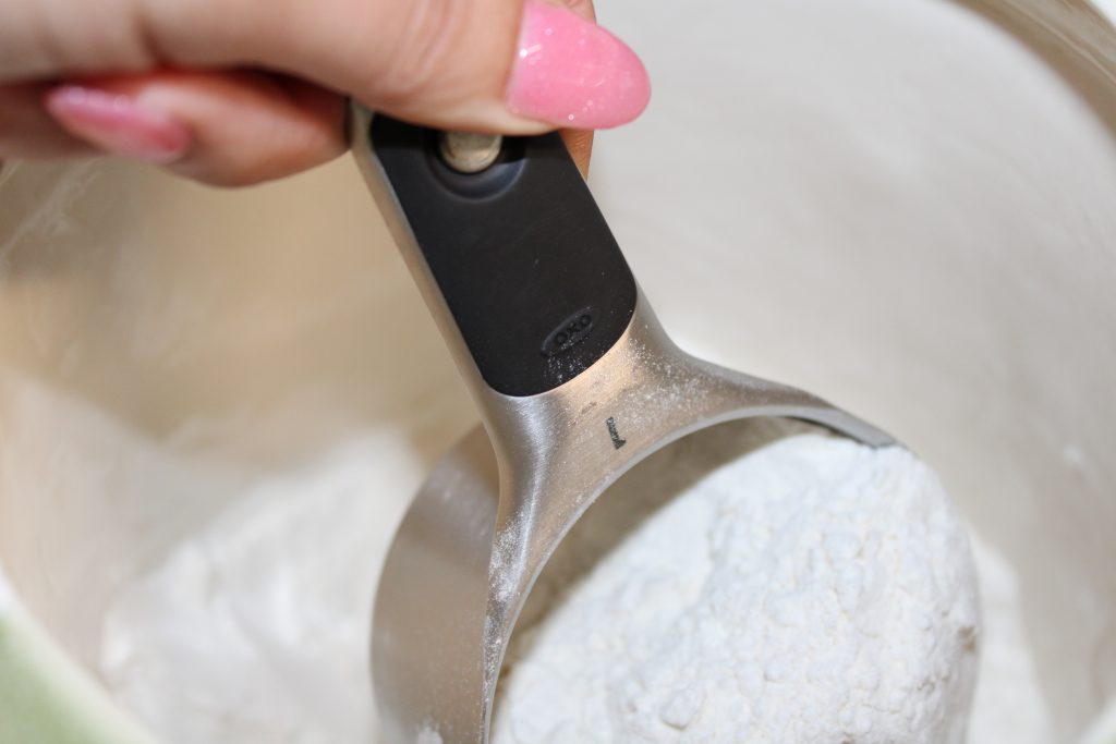 manicured fingers holding a measuring cup full of flour