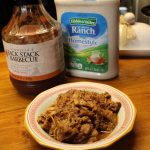 Pulled pork in a finishing sauce in a bowl next to a bottle of ranch and a bottle of barbecue sauce